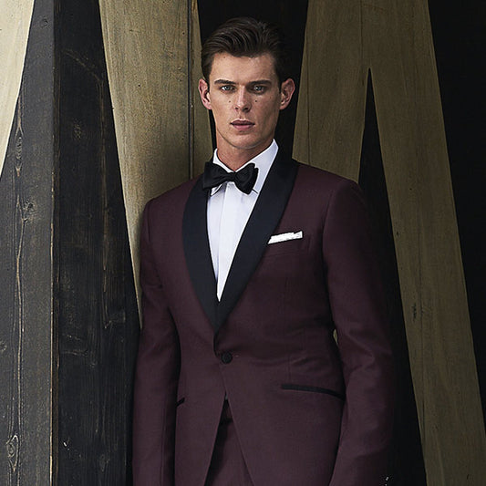 man wearing burgundy tuxedo with black bowtie stadning against a dark wood background