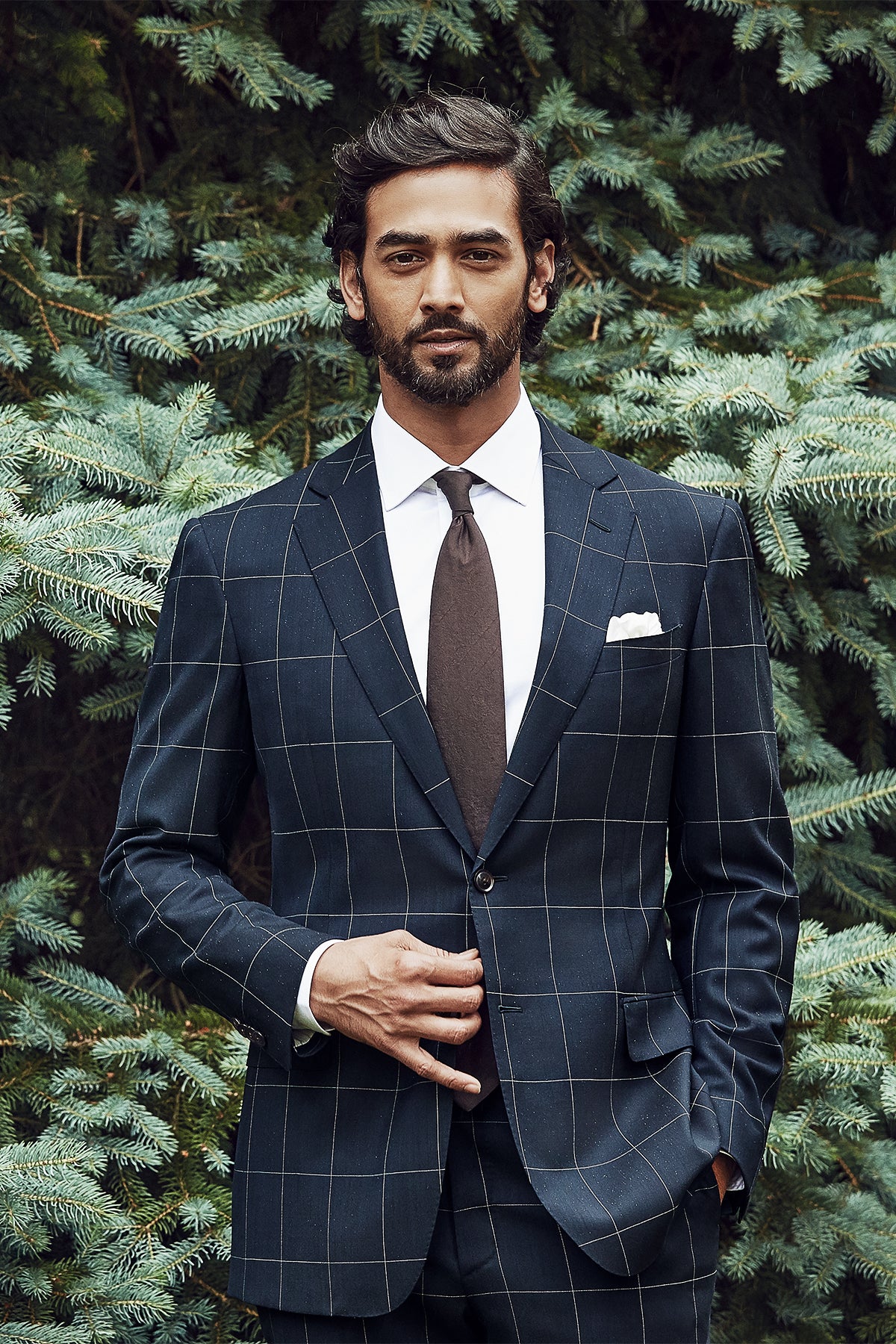 Man in a dark suit with a patterned tie standing in front of green foliage